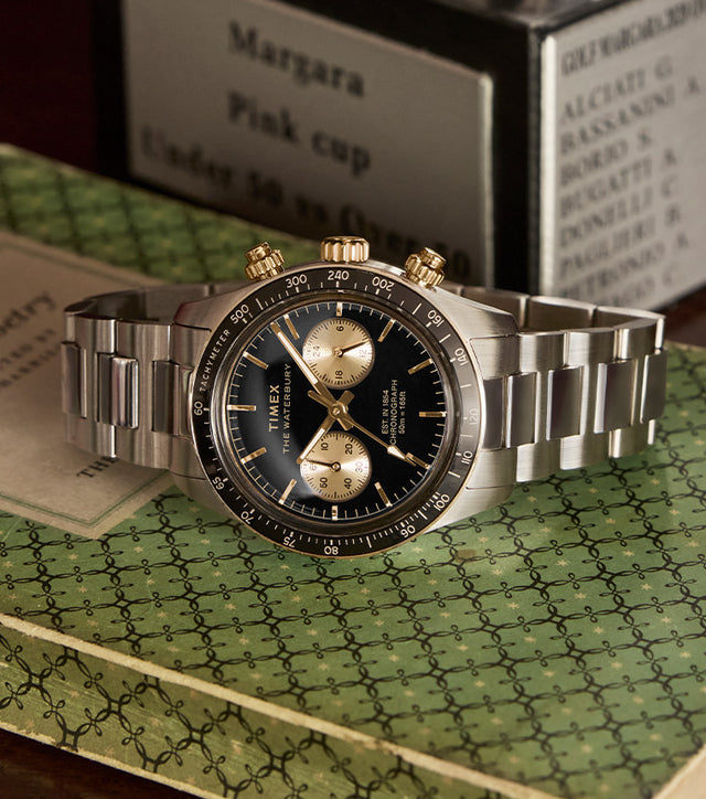 A close-up shot of the Timex Waterbury Heritage Chronograph resting on a vintage green-patterned book. The stainless steel bracelet, black dial with gold subdials, and gold-tone pushers and crown stand out, evoking a classic and refined aesthetic.