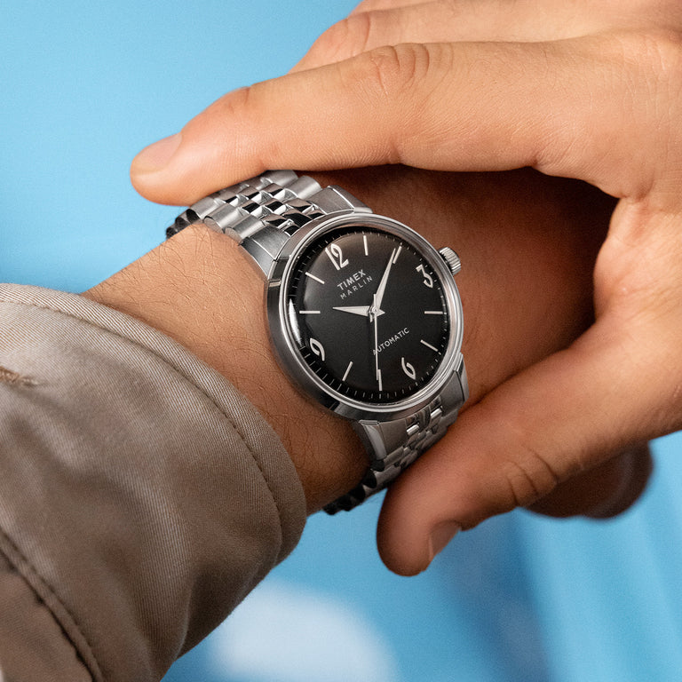 Timex Marlin automatic watch with a black dial, silver-tone stainless steel case and bracelet, worn on a wrist against a light blue background.