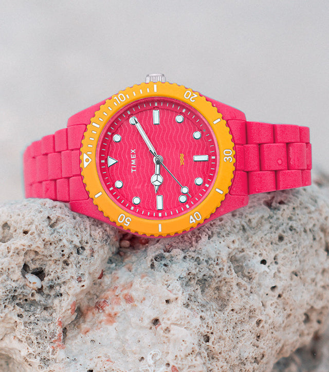 A vibrant pink and orange Timex Legacy #tide™ watch placed on a textured rock at the beach, highlighting its bold design and ocean-inspired theme.
