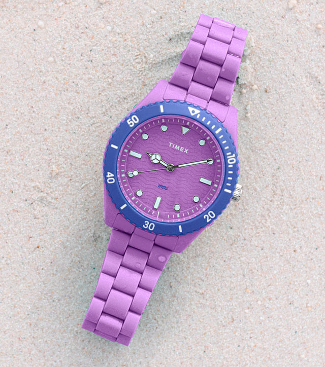 A purple Timex Legacy #tide™ watch with a blue bezel rests on soft sand near a coral rock, showcasing its eco-friendly design made from upcycled ocean-bound plastic.