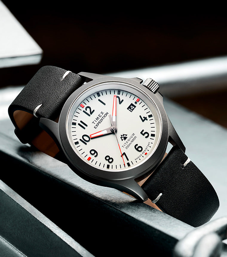 Timex Expedition Titanium watch featuring a lightweight titanium case, white dial, black leather strap, date display, and 200m water resistance, styled on an industrial metal background.
