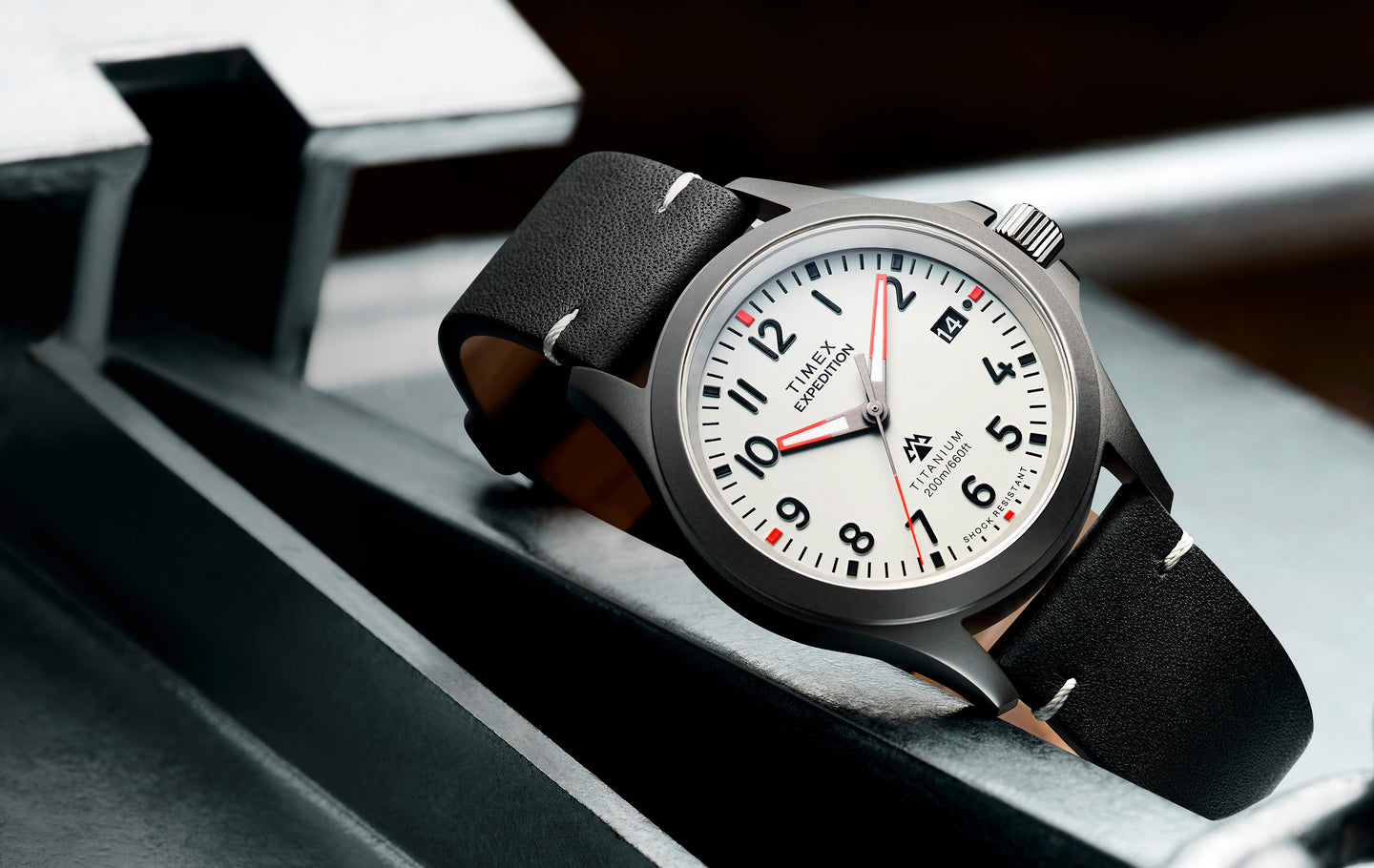 Timex Expedition Titanium watch featuring a lightweight titanium case, white dial, black leather strap, date display, and 200m water resistance, styled on an industrial metal background.
