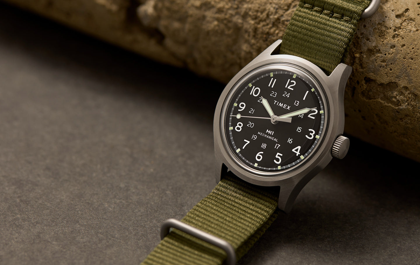 Timex® MK1® Hand-Wound watch with a brushed gray case, black field dial, and olive green fabric strap, displayed on a stone surface.