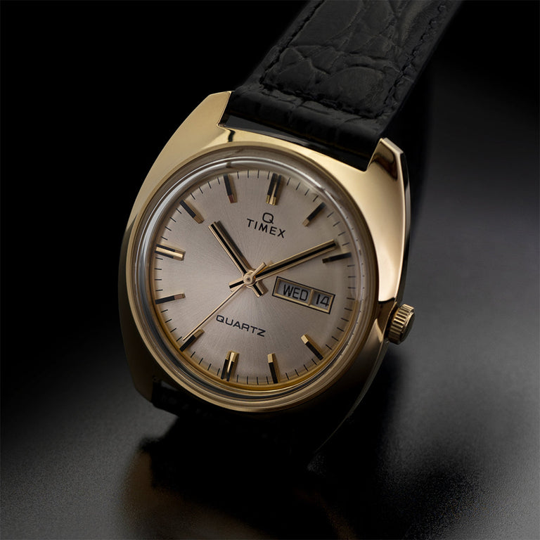 A gold-tone Timex Q Reissue with a champagne dial and black leather strap. The dial features baton hour markers and day-date display, photographed on a dark, moody surface to emphasize elegance.