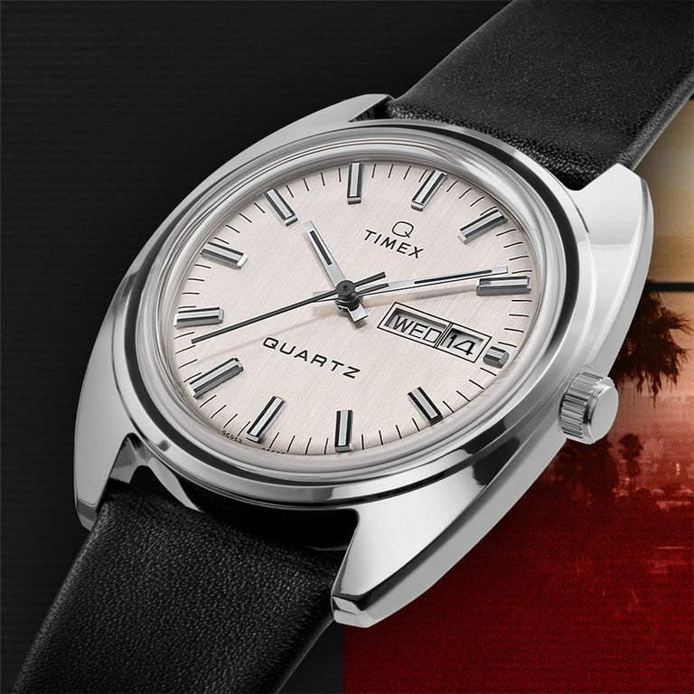 Silver-tone Timex Q Reissue watch with a brushed silver dial and black leather strap. Day-date window at 3 o'clock. The background features a cinematic dusk-toned gradient and palm tree silhouette.