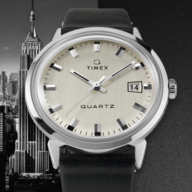 Timex Q Reissue watch with brushed champagne dial, minimal date window, and silver-tone hands and markers. Set against a black-and-white backdrop of the Empire State Building and city skyline.