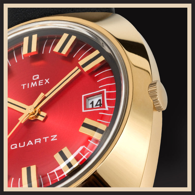 Bold Timex Q Reissue with a red sunburst dial, gold-tone hands and indices, and a gold case. Date window at 3 o'clock. The close-up reveals the brushed texture and polished lugs on a dark background with a beige border.