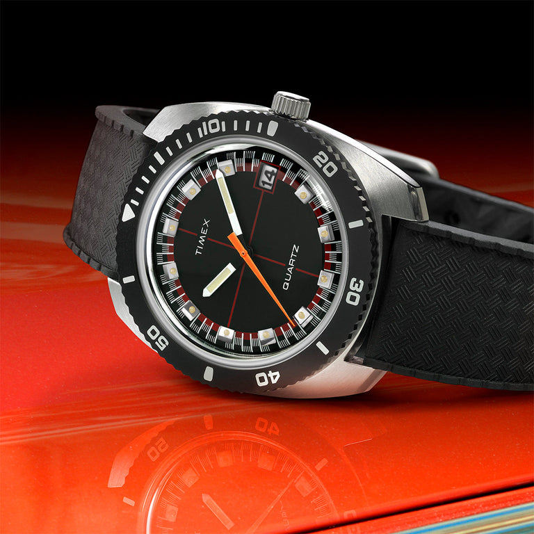 Diver-style Timex Q Reissue watch with black dial, orange seconds hand, and rotating black bezel. Features a textured black strap and is dramatically placed on a reflective red surface for a sporty appeal.