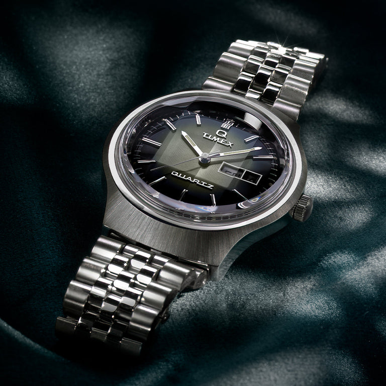 Timex Q Reissue watch featuring a brushed silver-tone case, a sunburst green dial with gradient fading to black, and a stainless steel bracelet. The watch is photographed on a dark green satin background, highlighting its elegant retro-inspired styling.