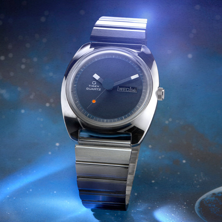 Timex Q Reissue watch with a minimalist navy blue dial, a bright orange seconds marker dot, and silver-tone hands and indices. The polished case and wide-link bracelet give it a futuristic aesthetic, shown against a moody, cosmic blue backdrop.