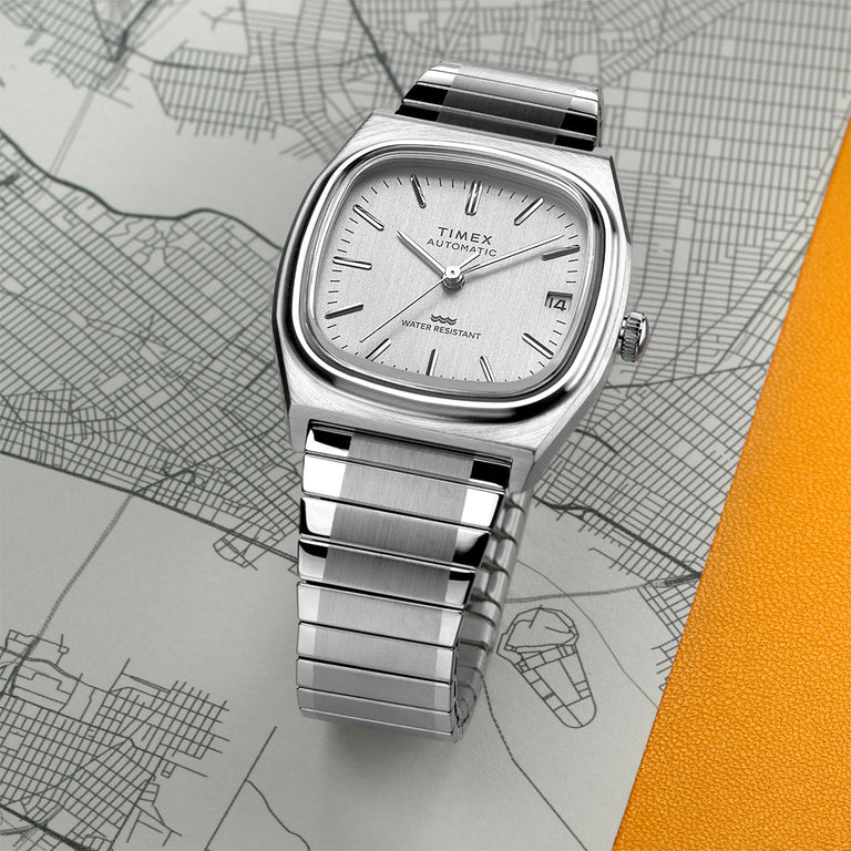 Timex M79 Automatic watch in stainless steel with a silver-tone cushion-shaped case and bracelet. The dial is minimalist with baton indices and date window at 3 o'clock. The watch rests on a city map illustration with a pop of orange texture for contrast.