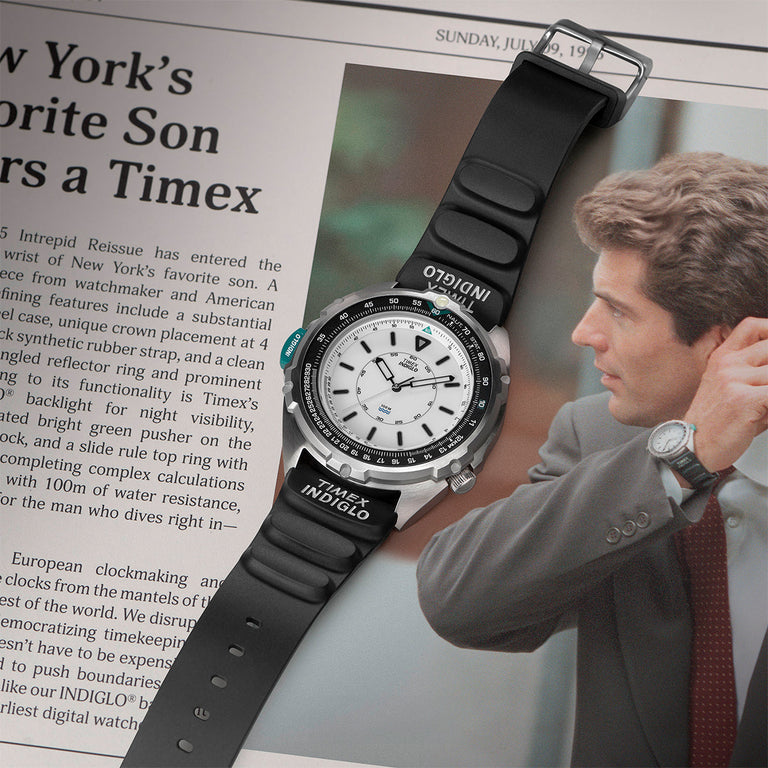 Timex Intrepid Reissue watch with white dial, black bezel, and black rubber strap featuring the INDIGLO® logo. Laid on top of a newspaper article with a photo of JFK Jr. wearing a similar model, emphasizing its nostalgic American icon status.