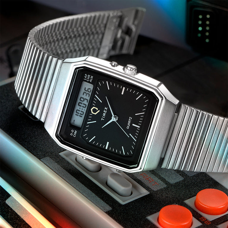 Timex Q Reissue digital-analog combo watch with a sleek black dial, rectangular digital display, and stainless steel expansion band. Placed atop a retro video game console controller, emphasizing its '80s-inspired tech design.