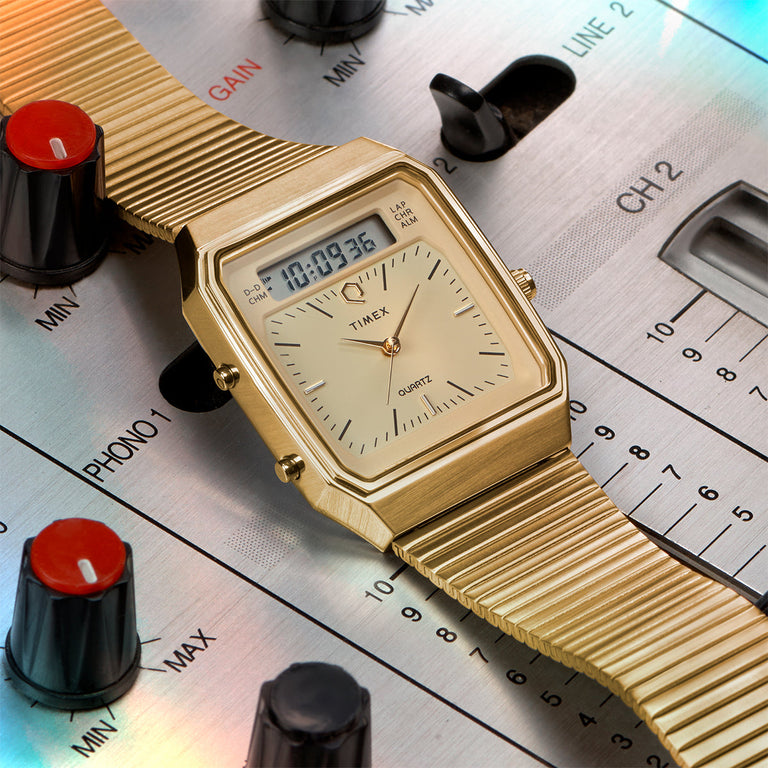 Gold-tone Timex Q Reissue watch with champagne dial, rectangular digital display, and slim expansion band. Positioned on a professional DJ sound mixer, reflecting its retro-futuristic flair with music-inspired styling.