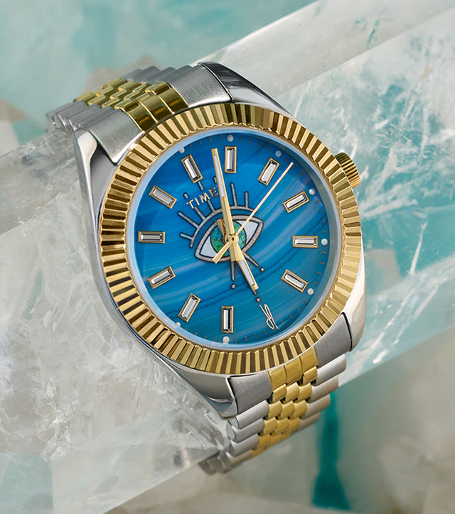 Timex x Jacquie Aiche watch resting on clear quartz crystals. The watch features a two-tone gold and silver bracelet, a fluted gold bezel, and a vibrant turquoise dial with a central eye motif embellished with gemstone detailing and radiant lines extending outward.