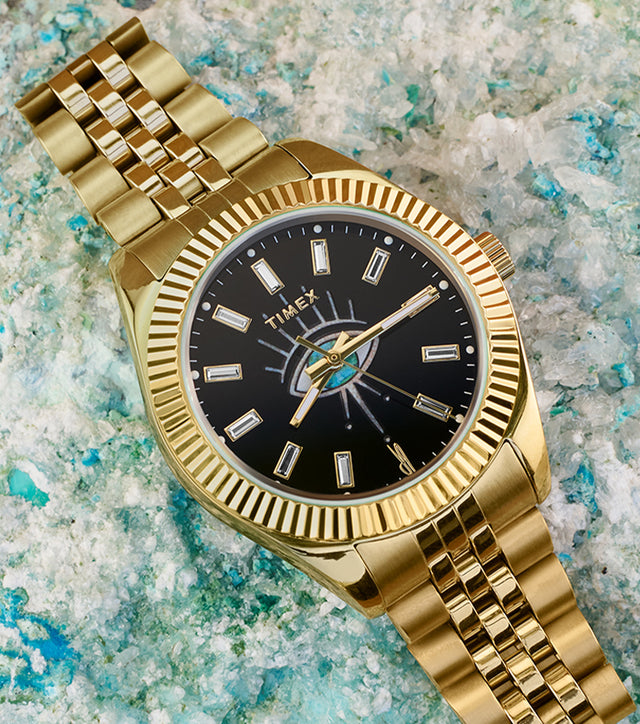 Timex x Jacquie Aiche gold-tone watch placed on textured blue and white mineral crystals. The timepiece features a polished gold bracelet and case, a fluted bezel, and a striking black dial with a central eye motif and baguette crystal hour markers.
