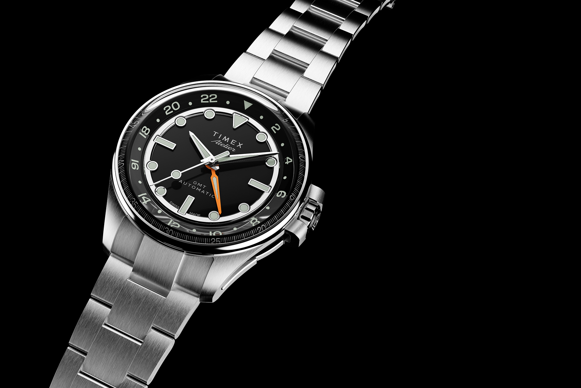 A Timex Atelier GMT Automatic watch with a black dial, orange GMT hand, and stainless steel bracelet displayed against a black background.