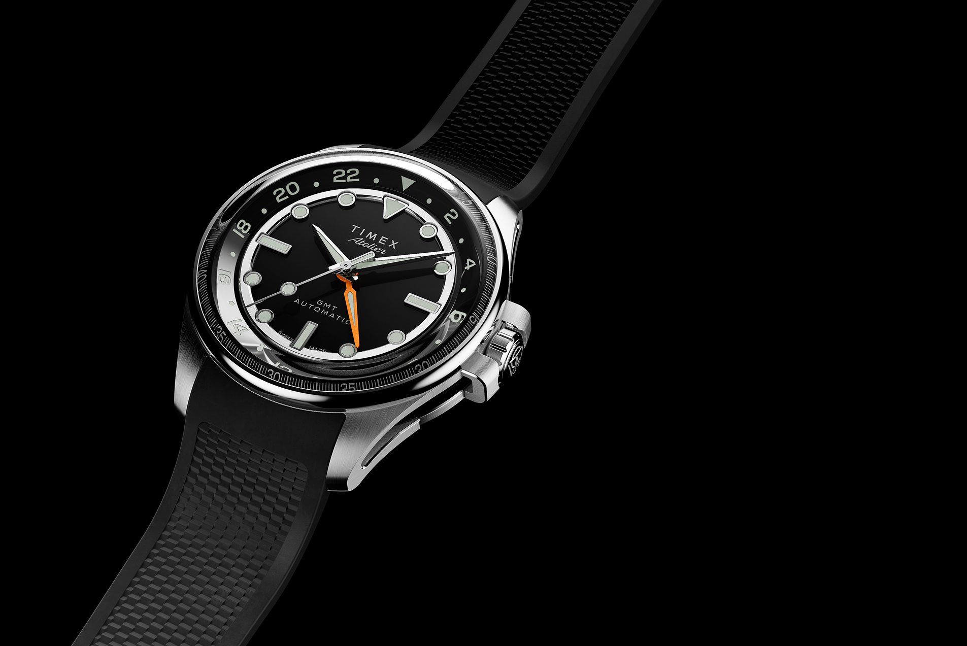 Timex Atelier GMT Automatic watch with a black dial, orange GMT hand, and black rubber strap displayed against a black background.
