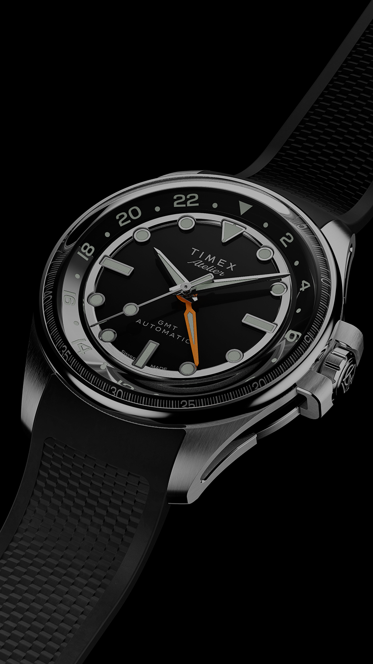 The Timex Atelier GMT Automatic watch with a black dial, orange GMT hand, and black rubber strap displayed against a black background.