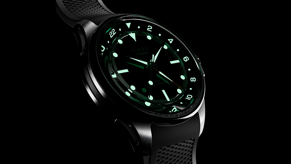 The Timex Atelier GMT Automatic glowing in the dark, displaying luminous green hour markers and hands.