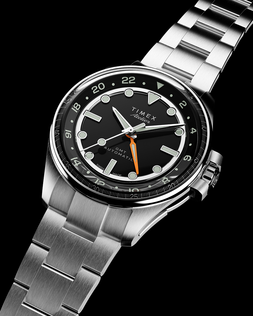 The Timex Atelier GMT Automatic with a stainless steel bracelet and black dial, featuring a bold orange GMT hand, shot from an angled perspective.