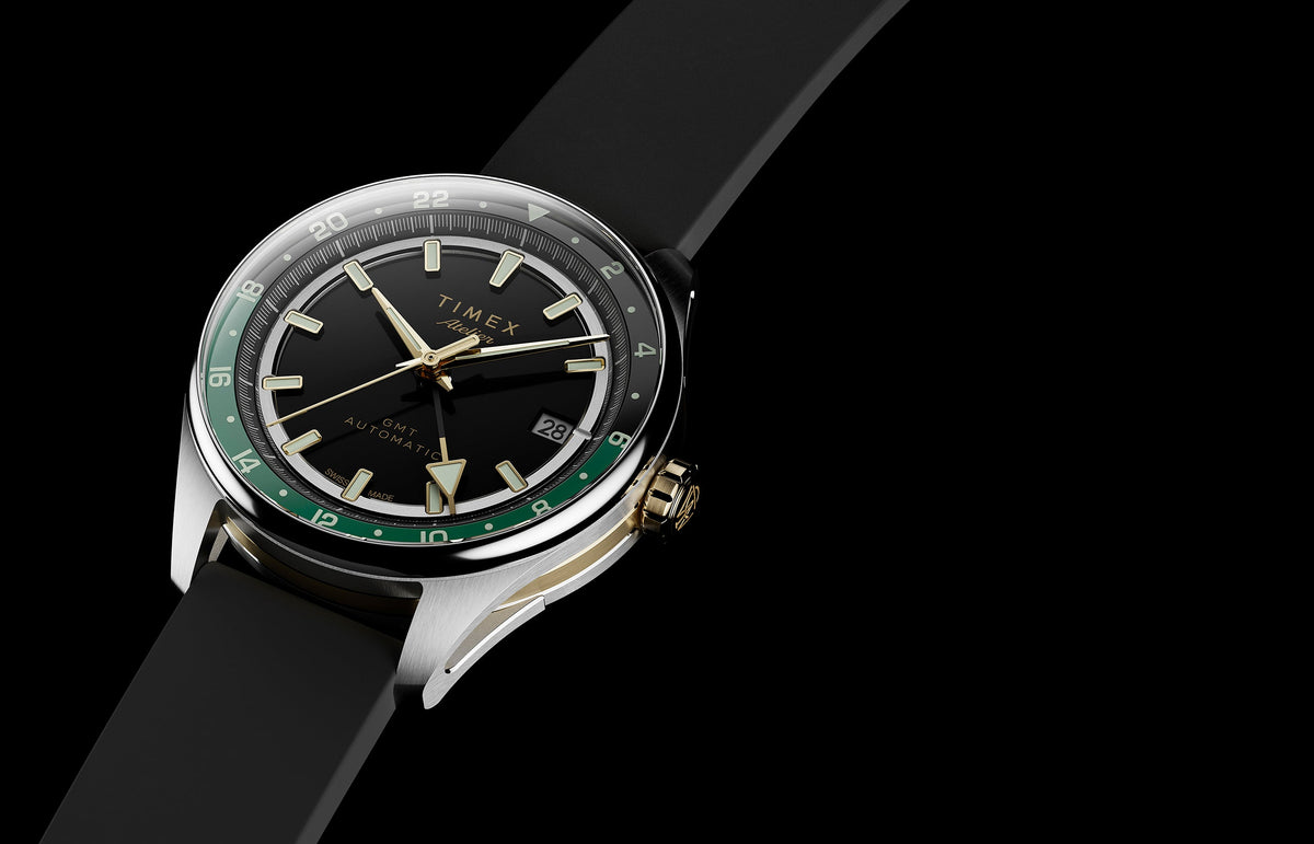 Timex Atelier GMT Première M1a watch with a black dial, green 24-hour scale, gold-tone accents, and black rubber strap.