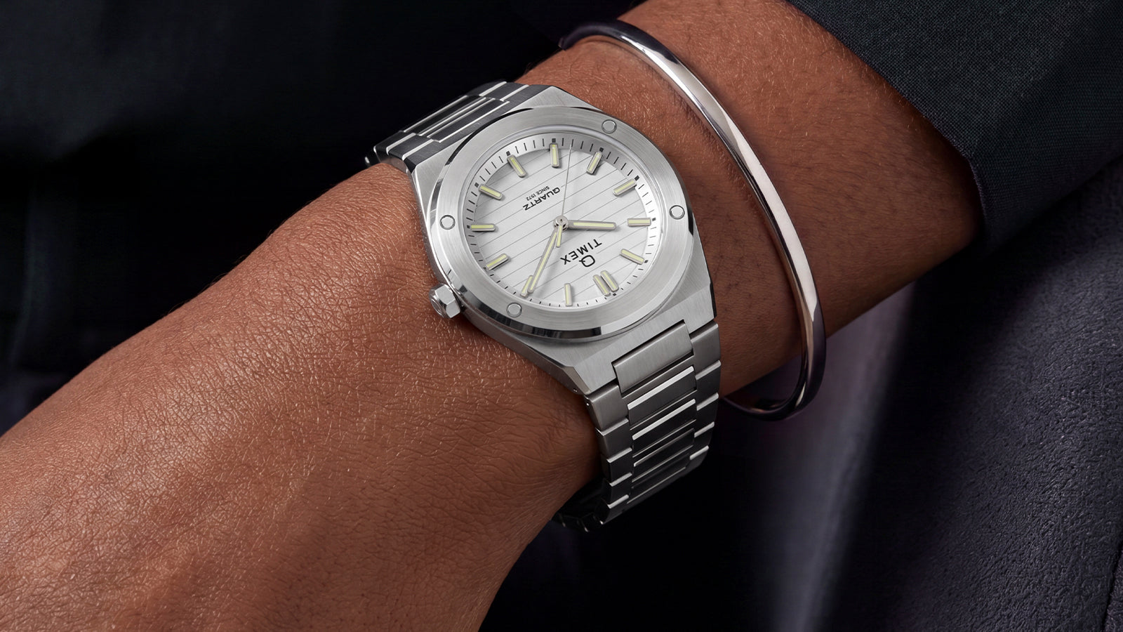 Q Timex Marbella watch with a silver-tone stainless steel case and integrated bracelet, featuring a silver sunray dial with luminous hands and markers, worn on the wrist with a silver bangle.