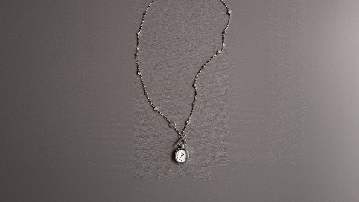 TW6A17600 Cavatina® Mini stainless steel necklace with a watch pendant, toggle clasp, and chain with clear stones, laid on gray fabric.