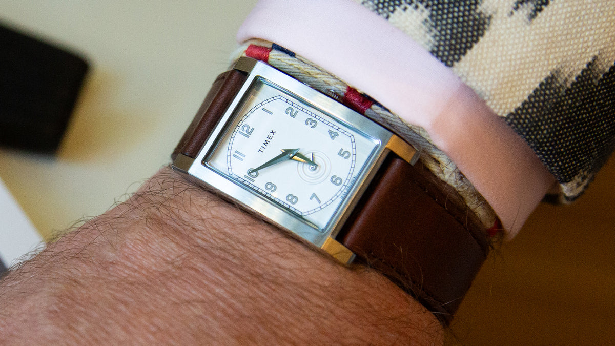 TWG069000 Close-up of a WM Brown x Timex rectangular watch with a brown leather strap, worn below a pink cuff and patterned jacket.