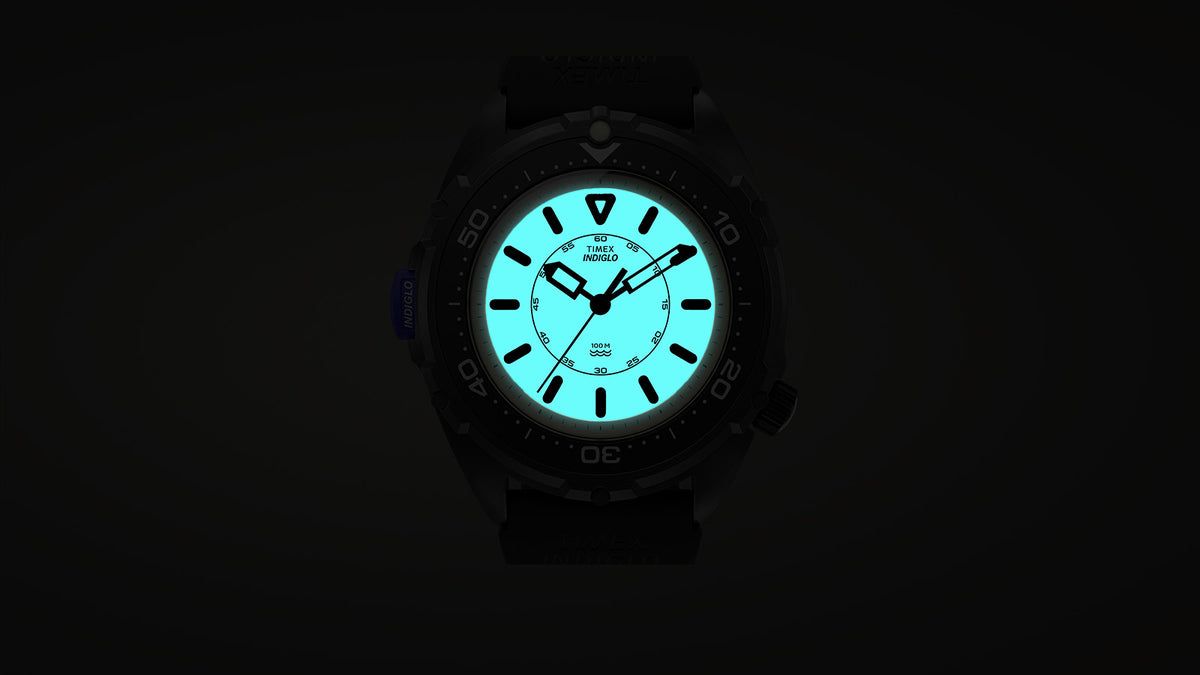 TW6A19800 Timex Intrepid x Dimepiece ‘Blackout’ Edition with its dial illuminated turquoise by the INDIGLO backlight against a dark background.