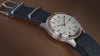 Marlin Jet America 250 automatic watch with a white dial, red bezel accent, red seconds hand, and blue woven strap displayed on a reflective blue surface.